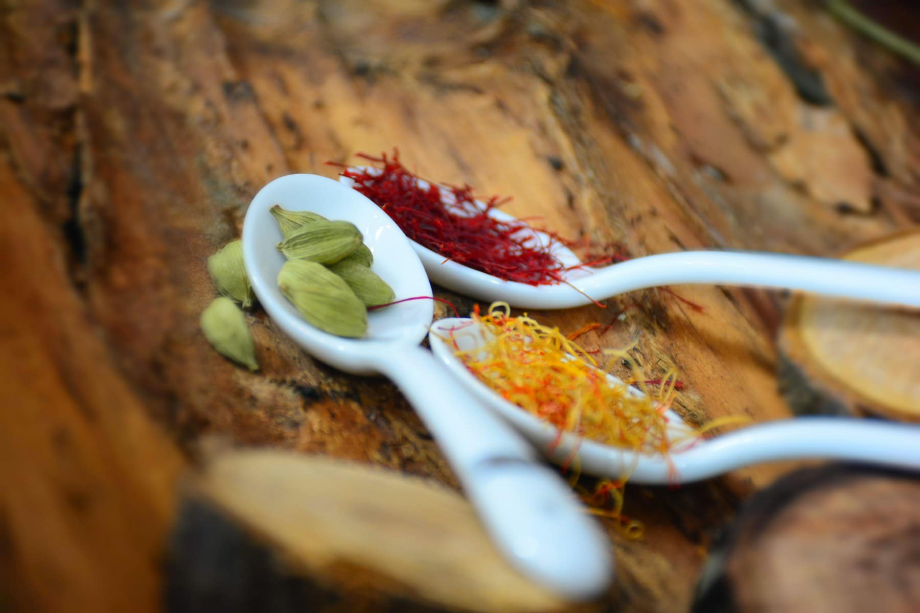 Saffron threads and green cardamom pods in white ceramic spoons on dark wood
